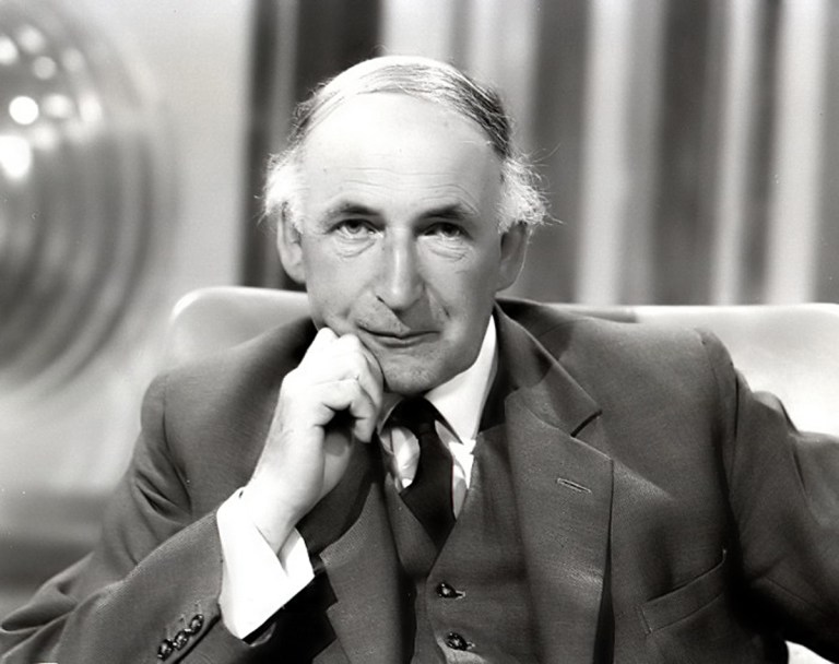 A black-and-white portrait features a man in a three-piece suit and tie seated in the center frame, resting his chin on his right hand while looking directly forward. Bright studio lighting illuminates his face and light-colored shirt cuffs, creating smooth contrast against his dark jacket and vest. The background consists of soft-focus, vertically striped curtains and a blurred metallic sphere to the left.