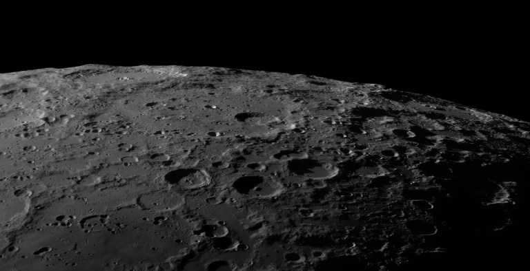 A jagged, heavily cratered lunar landscape curves along the horizon, lit at a low, raking angle so every crater rim and ridge casts a long, sharp shadow. Countless overlapping craters of all sizes cover the gray surface, fading into darkness where the Moon's curve meets black sky.