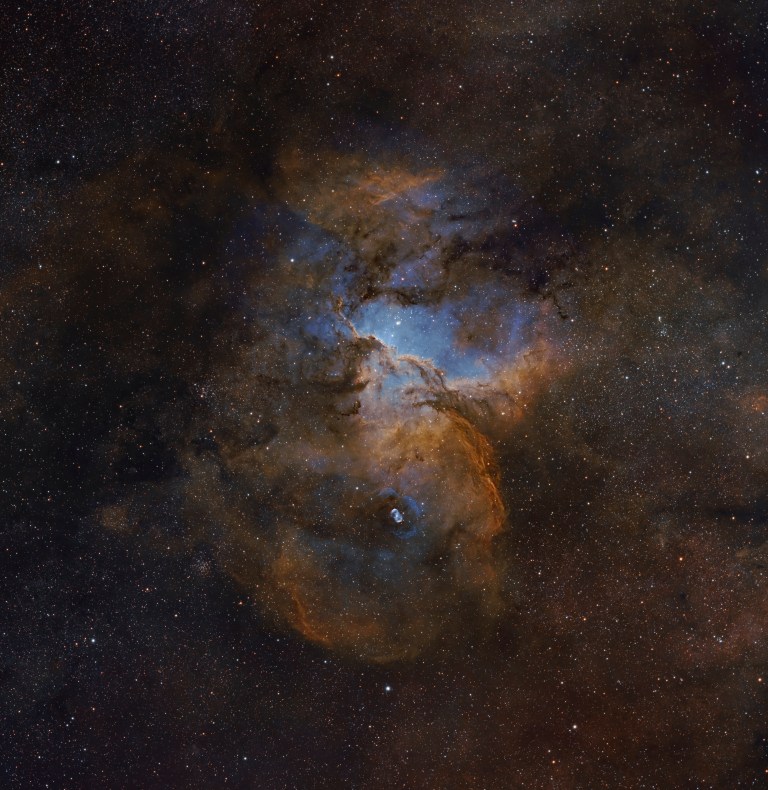 A dark, dust-laced cloud glows red and orange against a dense starfield, with pale blue light seeping through gaps near its center and top. Near the bottom, a small, distinct blue-and-white oval nebula glows within the surrounding dust.