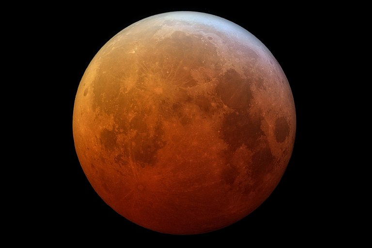 A detailed telescopic photograph of a total lunar eclipse, showing the Moon glowing in rich shades of deep red, orange, and brown — a blood moon. The Moon's surface features are clearly visible, including craters, ray systems, and dark maria regions. A sliver of brighter white-gray light is visible along the upper edge, indicating the Moon is partially outside Earth's umbral shadow.