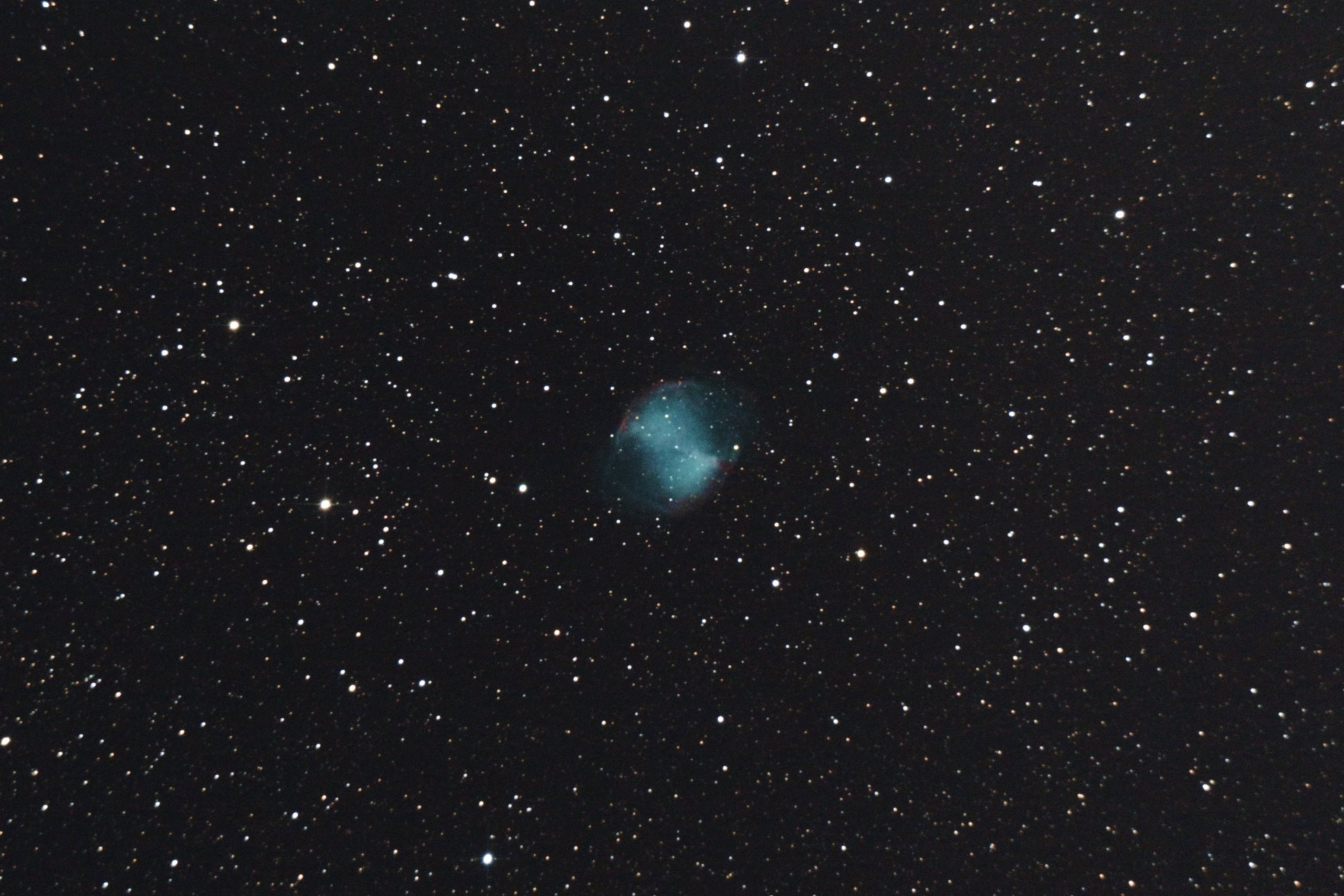 A telescopic photograph of M27, the Dumbbell Nebula, showing the planetary nebula's distinctive double-lobed shape glowing in teal-blue with hints of red at the pinched edges, set against a black sky scattered with orange and white foreground stars.