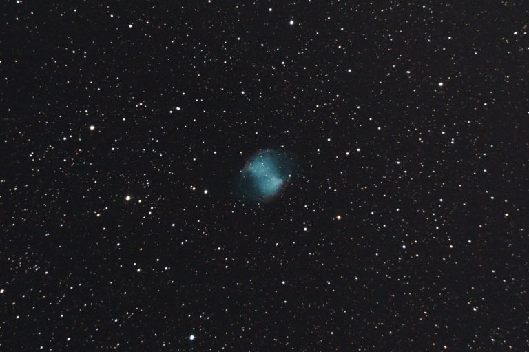A telescopic photograph of M27, the Dumbbell Nebula, showing the planetary nebula's distinctive double-lobed shape glowing in teal-blue with hints of red at the pinched edges, set against a black sky scattered with orange and white foreground stars.