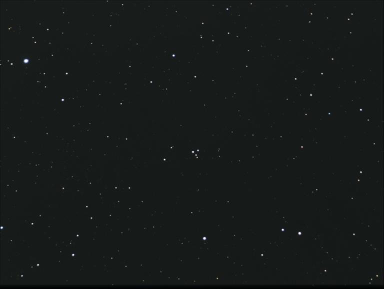 A telescopic photograph showing M73, a small asterism of four stars arranged in a tight Y-shaped grouping near the center of the image, set against a sparse star field of faint orange and white points on a black background.