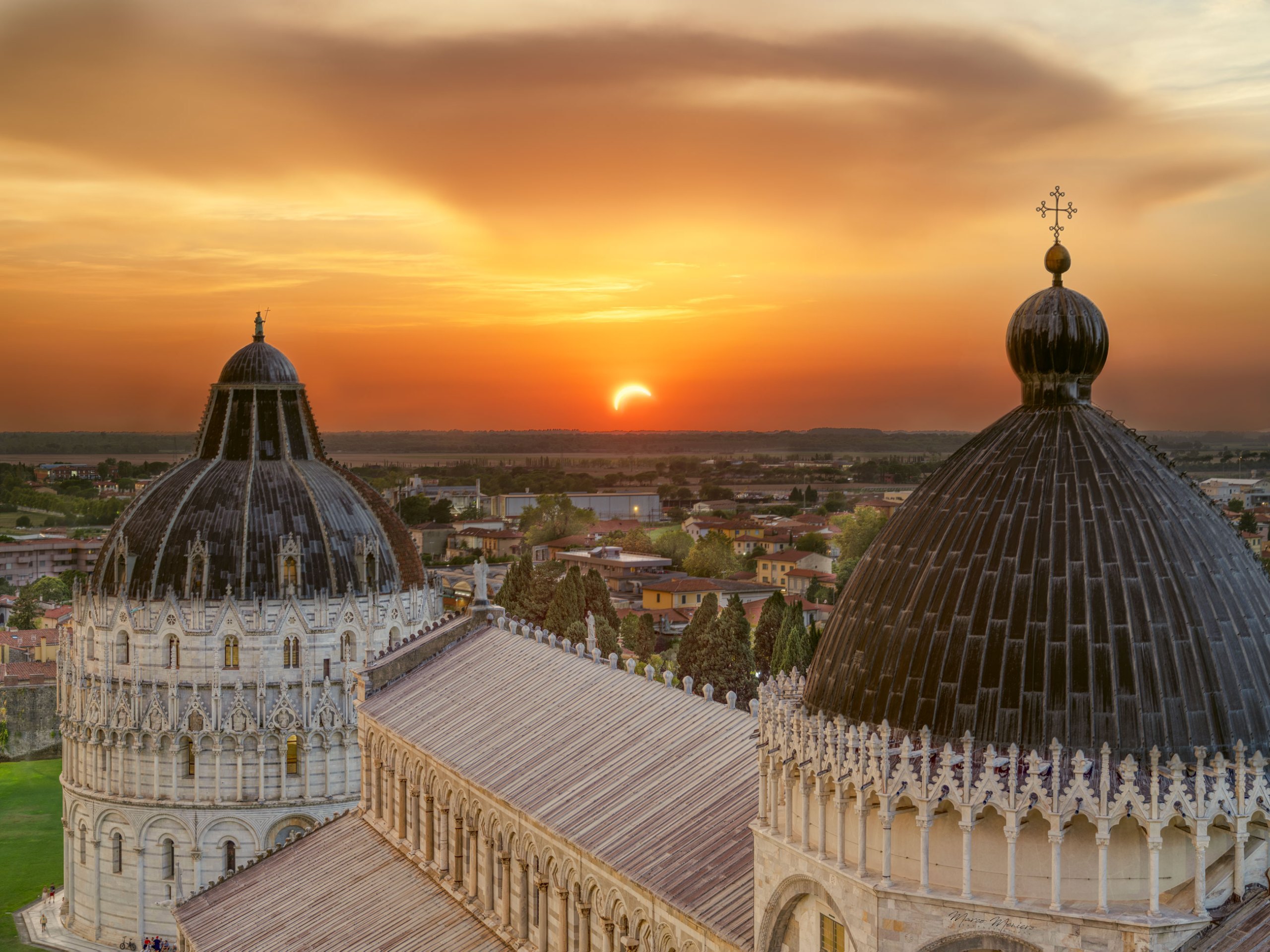 Two dark, ornately ribbed domes fill the lower half of the frame, their stone facades and columned arcades catching the last daylight. Above them, a thin crescent Sun glows low on the horizon. Above it, clouds streak across a deep orange and gold sky. In the background, a hazy patchwork of rooftops and trees stretch toward the horizon.