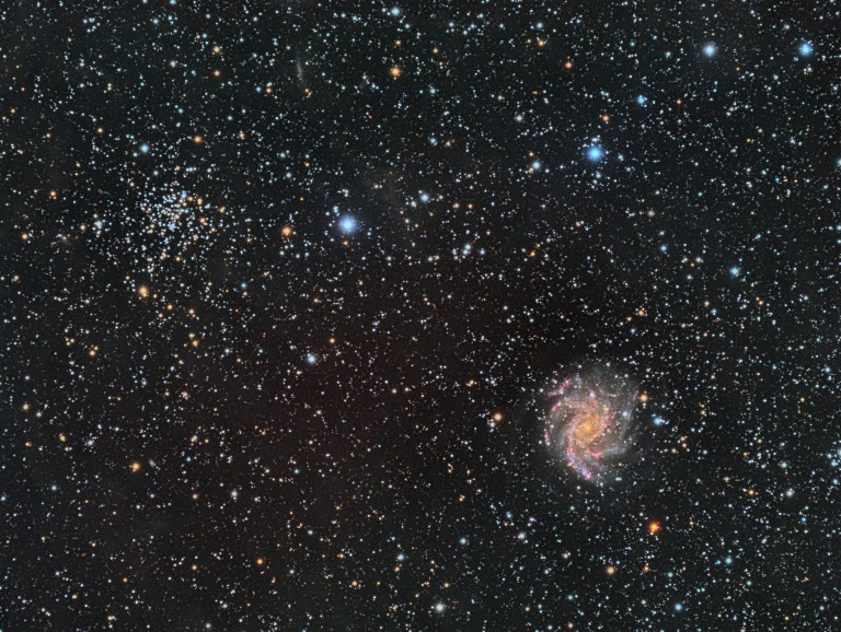 A telescopic photograph showing two objects side by side against a dense, colorful star field: NGC 6939, a rich open star cluster with a mix of blue-white and orange stars in the upper left, and NGC 6946, the Fireworks Galaxy, a face-on spiral galaxy with a golden core and pink-tinged spiral arms visible in the lower right.
