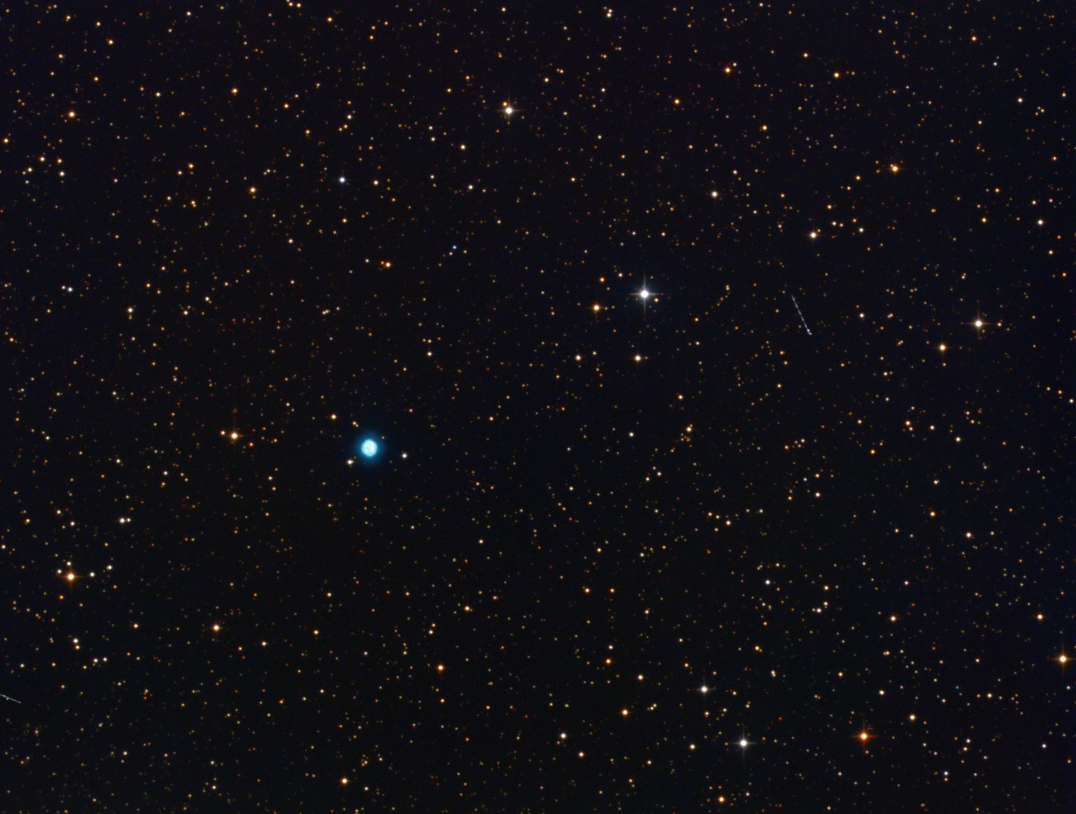 A telescopic photograph showing NGC 6818, the Little Gem Nebula, as a small, vivid teal-blue planetary nebula set among a field of orange and white foreground stars against a black sky. A faint satellite trail is visible as a short diagonal streak in the upper right portion of the image.
