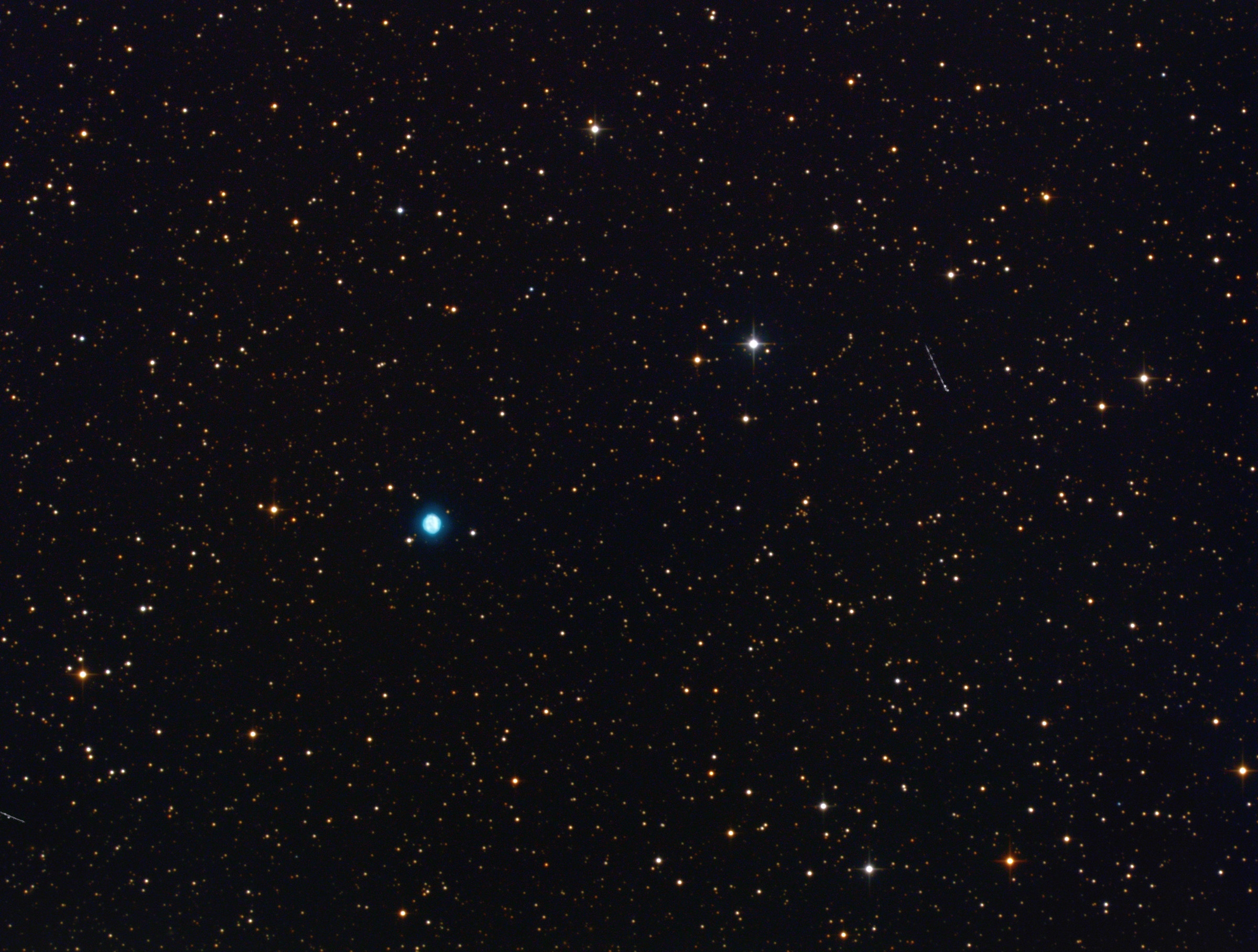 A telescopic photograph showing NGC 6818, the Little Gem Nebula, as a small, vivid teal-blue planetary nebula set among a field of orange and white foreground stars against a black sky. A faint satellite trail is visible as a short diagonal streak in the upper right portion of the image.