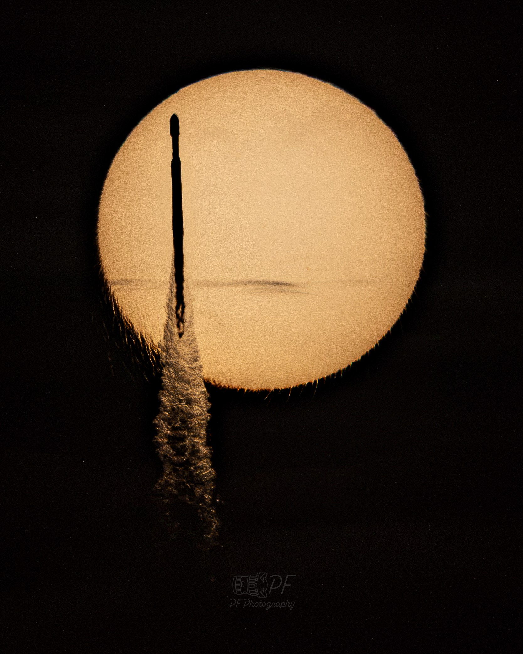 A large, glowing orange-tan disk — the Sun — fills most of the frame against a black sky. A thin, dark, needle-like silhouette crosses near its left edge, trailing a bright, feathery plume that distorts and refracts the image of the disk, extending below it. A faint dark smudge of clouds sits near the center of the disk, as well as small dark specks of sunspots.s