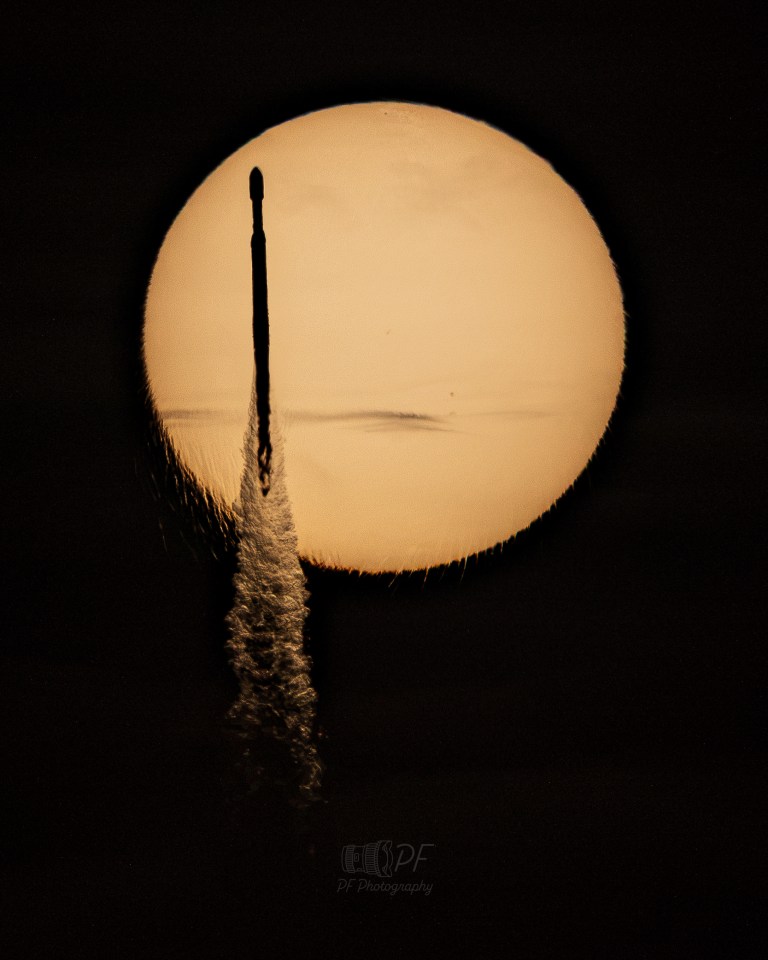 A large, glowing orange-tan disk — the Sun — fills most of the frame against a black sky. A thin, dark, needle-like silhouette crosses near its left edge, trailing a bright, feathery plume that distorts and refracts the image of the disk, extending below it. A faint dark smudge of clouds sits near the center of the disk, as well as small dark specks of sunspots.s