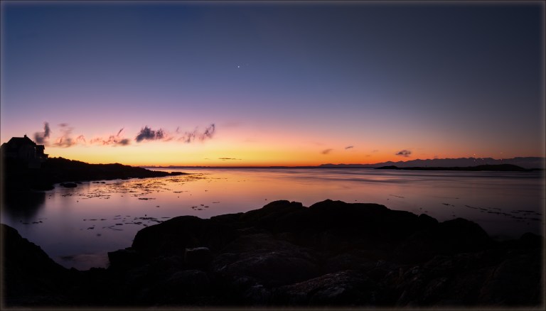 A panoramic twilight photograph taken along the rocky shore of the Salish Sea, looking west across calm water that mirrors the vivid sunset colors. Venus and Spica are visible as two close points of light in the deep blue sky above the center of the scene. The horizon glows in brilliant bands of orange and yellow, fading upward through purple to deep blue, with silhouetted clouds stretched across the middle sky. A silhouetted house is visible on the left, dark rocky outcroppings frame the foreground, and a distant mountain range is outlined against the orange glow on the right.