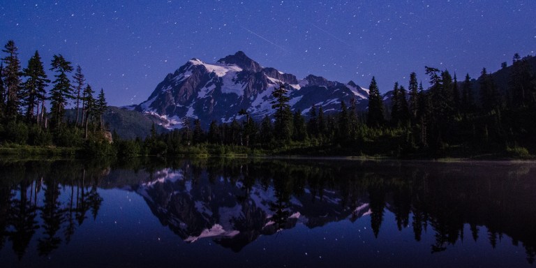 A nighttime landscape photograph of Mount Shuksan in Washington State, its glaciated rocky peak and surrounding evergreen trees perfectly reflected in the still waters of Picture Lake below. A Perseid meteor streaks diagonally across the deep blue starry sky above the summit. The scene is bathed in cool blue moonlight, with stars trailing slightly due to the long exposure.