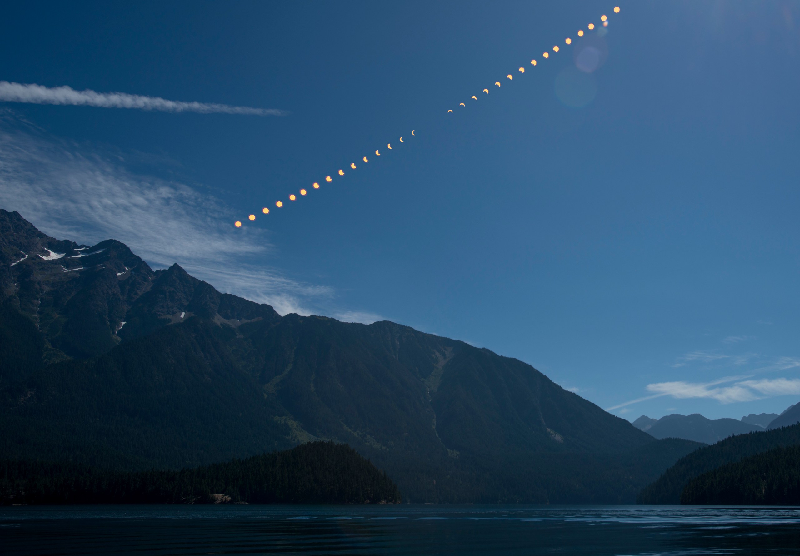 A composite photograph showing the progression of a total solar eclipse as a sequence of partially and fully eclipsed suns arcing diagonally across a blue sky above a mountainous landscape and calm lake. The phases progress from a crescent sun at lower left through totality near the middle, then back to a crescent at upper right.