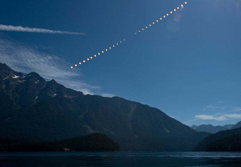 A composite photograph showing the progression of a total solar eclipse as a sequence of partially and fully eclipsed suns arcing diagonally across a blue sky above a mountainous landscape and calm lake. The phases progress from a crescent sun at lower left through totality near the middle, then back to a crescent at upper right.