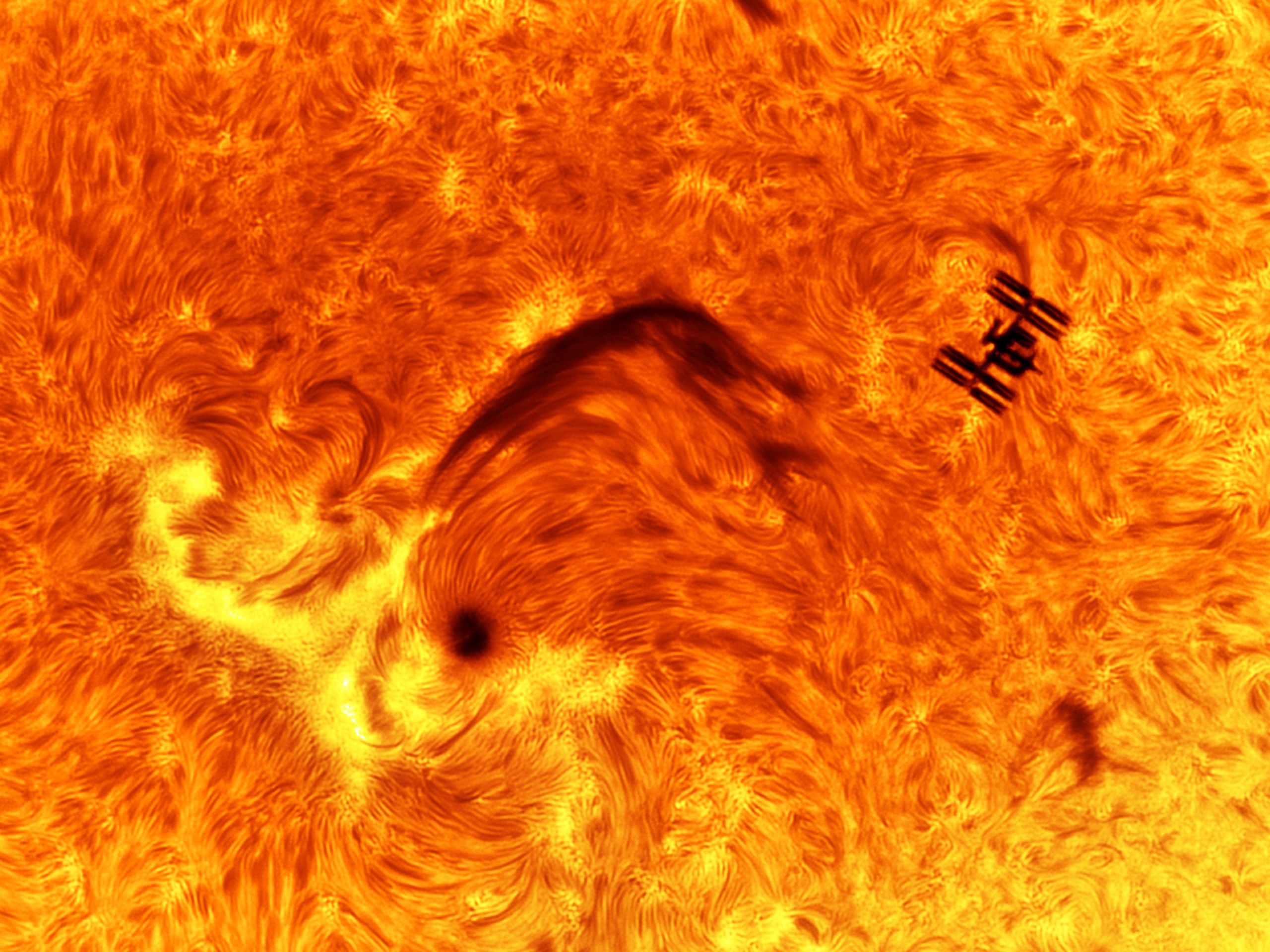 A close-up view of the Sun's surface shows a swirling, fibrous texture in deep orange and red, resembling flowing fur or fabric. A dark, arching loop of cooler gas curves across the center, and the tiny black silhouette of the space station, with its solar panels and modules visible, sits just to its upper right.