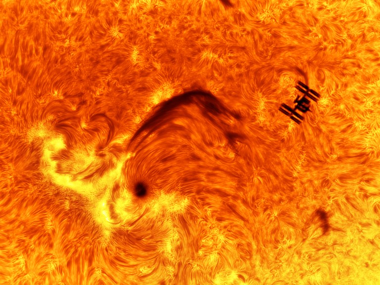 A close-up view of the Sun's surface shows a swirling, fibrous texture in deep orange and red, resembling flowing fur or fabric. A dark, arching loop of cooler gas curves across the center, and the tiny black silhouette of the space station, with its solar panels and modules visible, sits just to its upper right.