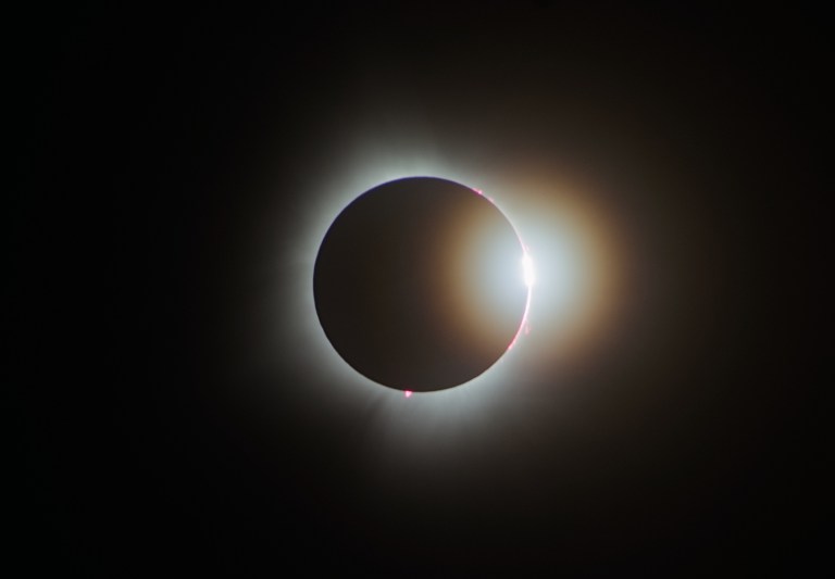 A photograph of a total solar eclipse capturing the diamond ring effect, where a single brilliant point of sunlight blazes on the right side of the Moon's silhouette just as totality ends, surrounded by the softly glowing white corona. Several pink solar prominences are visible along the Moon's edge, and a warm golden glow fans out around the right side of the disc against a black sky.