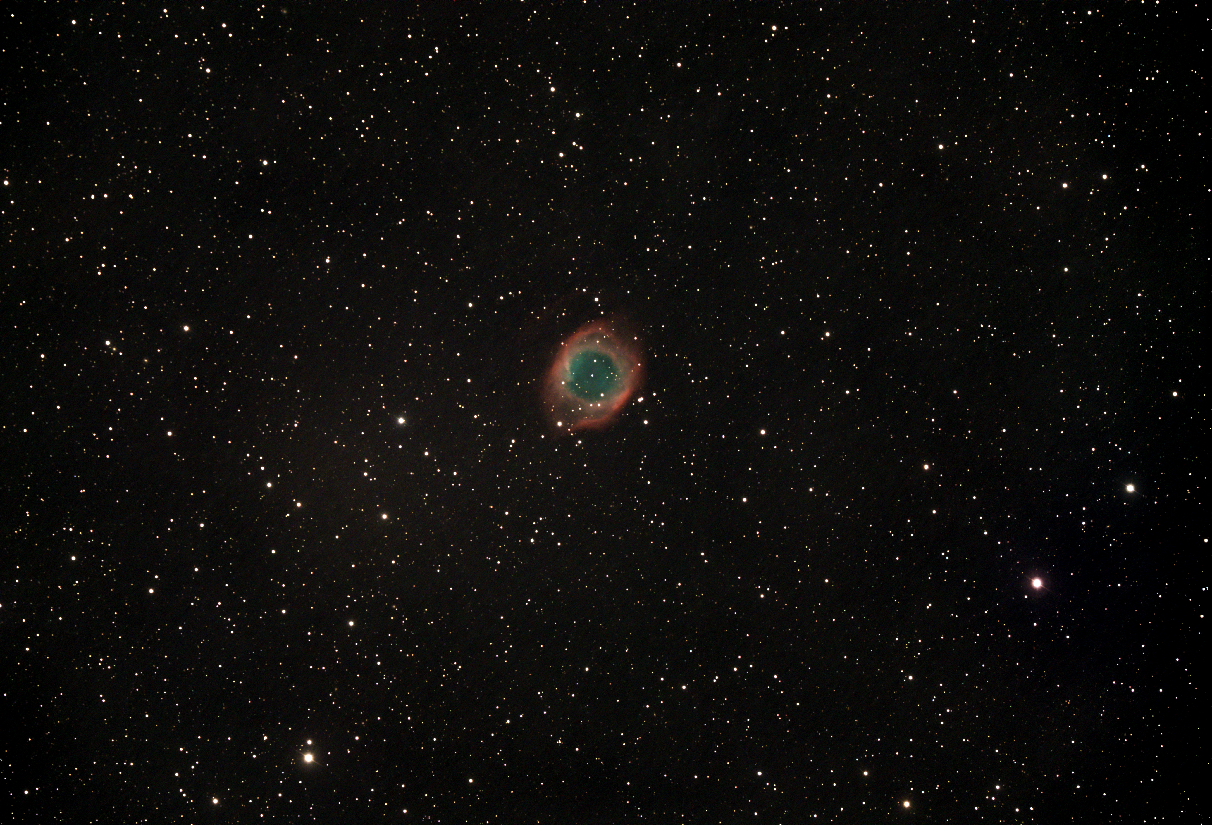 A telescopic photograph of the Helix Nebula (NGC 7293), a large planetary nebula appearing as a circular ring of glowing teal-green gas surrounded by a diffuse red outer halo, set against a black sky scattered with orange and white foreground stars.