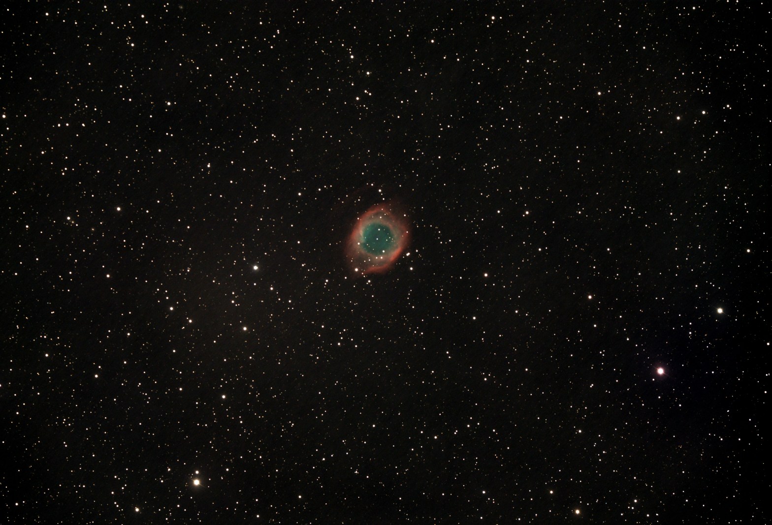 A telescopic photograph of the Helix Nebula (NGC 7293), a large planetary nebula appearing as a circular ring of glowing teal-green gas surrounded by a diffuse red outer halo, set against a black sky scattered with orange and white foreground stars.