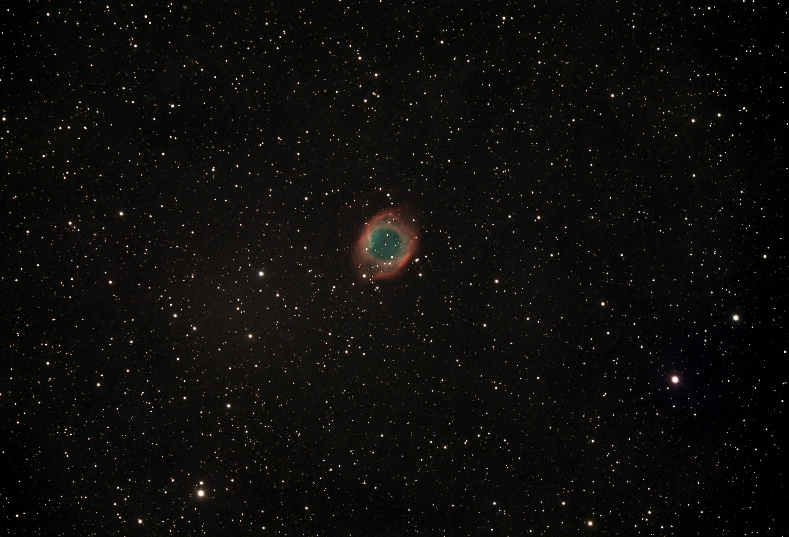 A telescopic photograph of the Helix Nebula (NGC 7293), a large planetary nebula appearing as a circular ring of glowing teal-green gas surrounded by a diffuse red outer halo, set against a black sky scattered with orange and white foreground stars.