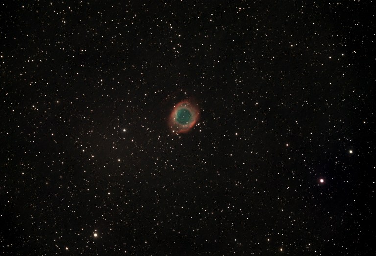A telescopic photograph of the Helix Nebula (NGC 7293), a large planetary nebula appearing as a circular ring of glowing teal-green gas surrounded by a diffuse red outer halo, set against a black sky scattered with orange and white foreground stars.