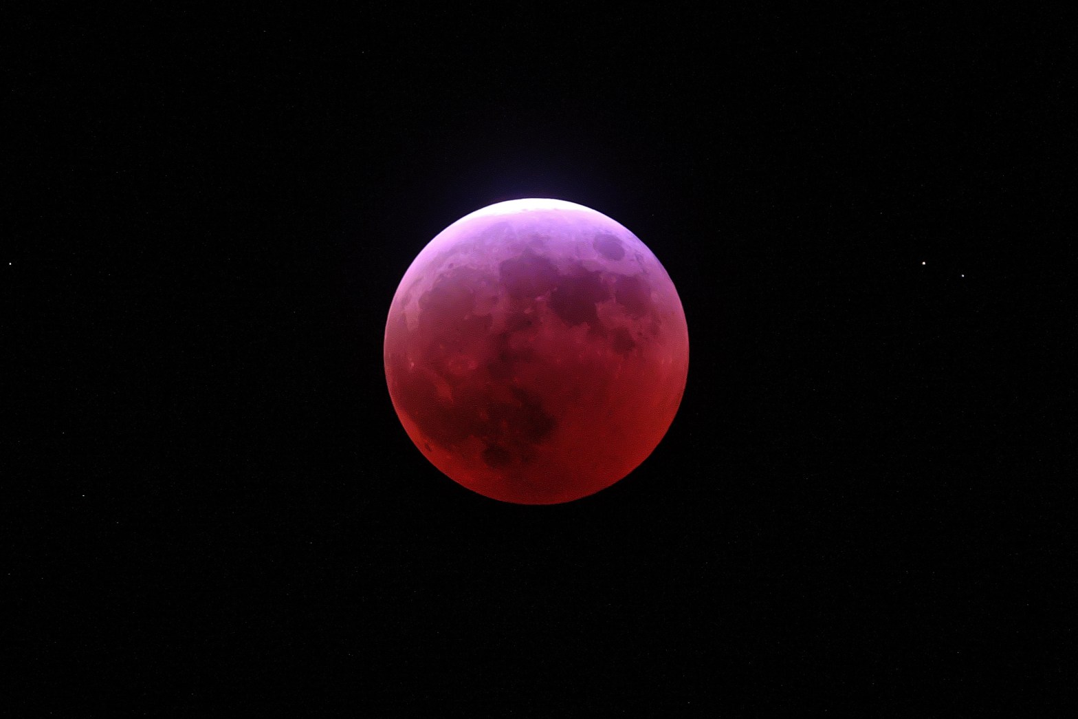 A photograph of a total lunar eclipse showing the Moon glowing in deep red and crimson hues — a blood moon — against a black sky. The Moon's surface features, including its darker maria regions, are still faintly visible through the red coloration. A thin sliver of brighter bluish-white light is visible along the upper edge, suggesting the Moon is just beginning to exit or enter Earth's umbral shadow.