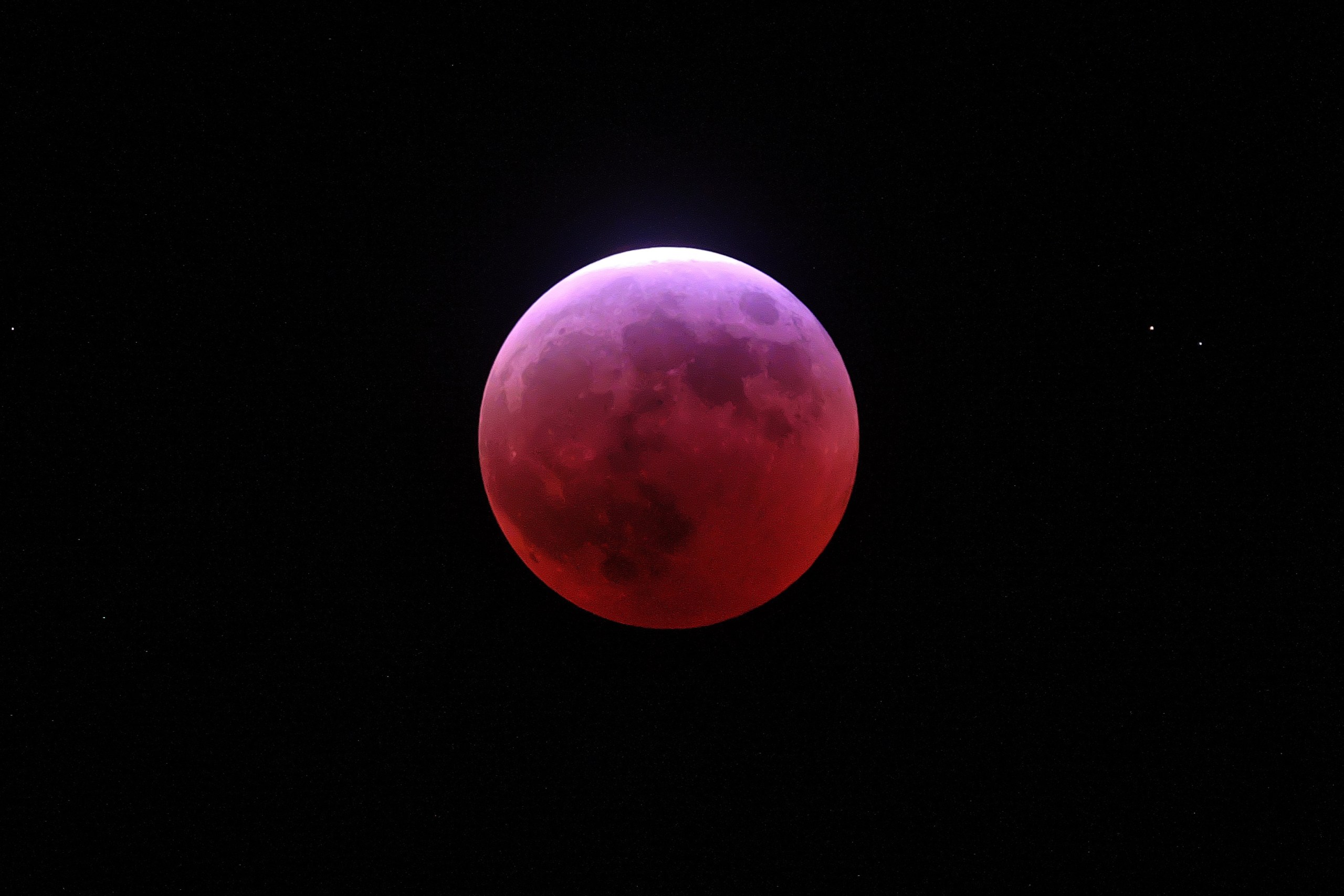A photograph of a total lunar eclipse showing the Moon glowing in deep red and crimson hues — a blood moon — against a black sky. The Moon's surface features, including its darker maria regions, are still faintly visible through the red coloration. A thin sliver of brighter bluish-white light is visible along the upper edge, suggesting the Moon is just beginning to exit or enter Earth's umbral shadow.