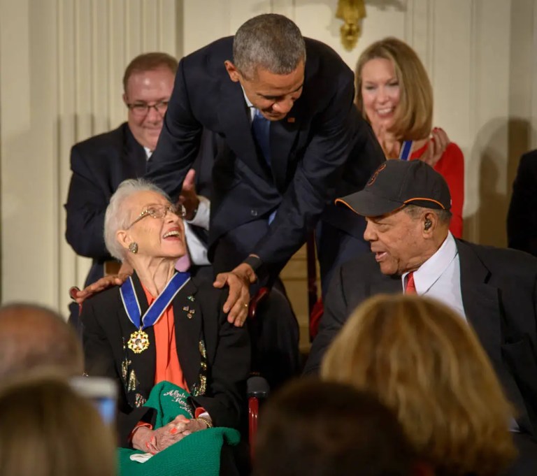 A medium shot shows a man in a dark suit leaning down with both hands resting gently on the shoulders of an elderly woman seated in the center foreground. The smiling woman wears a dark jacket, a bright orange shirt, and a gold medal around her neck attached to a blue ribbon, looking up toward the man. Other seated guests surround them against a cream-colored background, all under bright interior lighting that creates sharp detail throughout the frame.