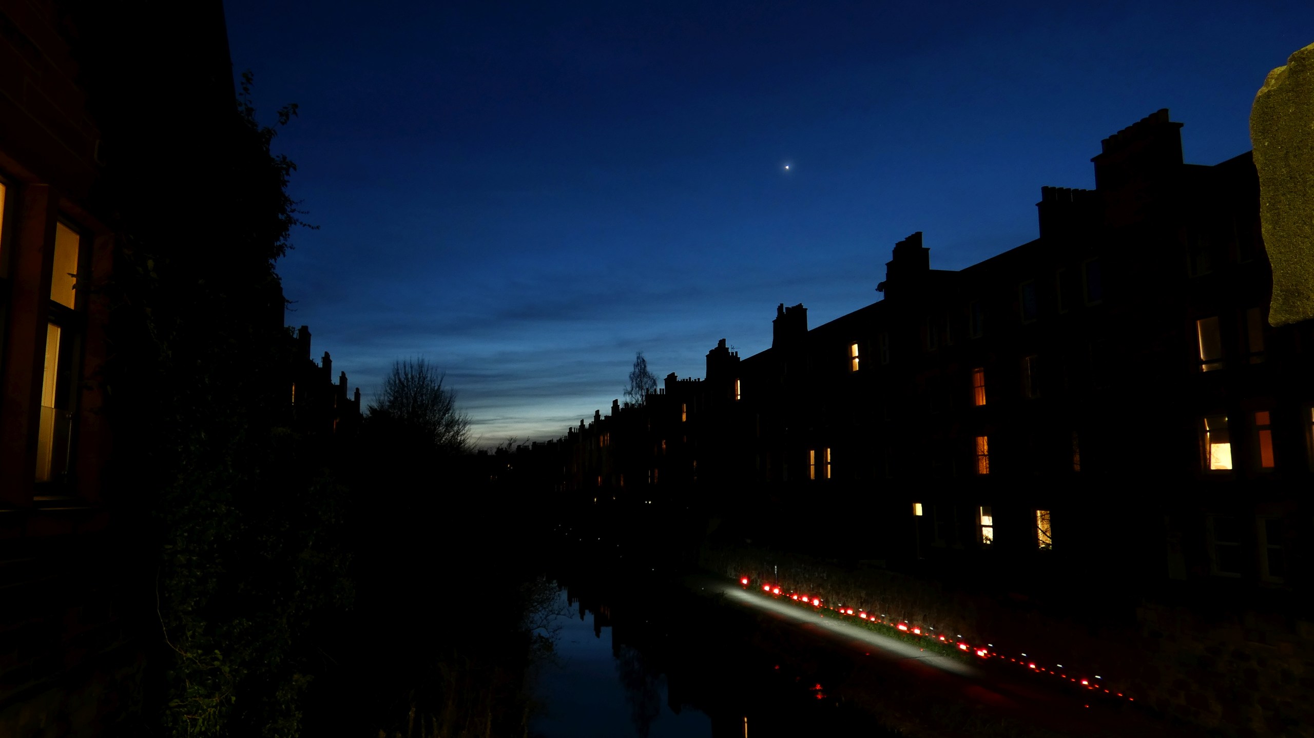 A twilight photograph taken along a narrow urban canal, with rows of silhouetted terraced buildings lining both sides and their lit windows glowing warm orange. Venus shines as a bright point of light in the deep blue evening sky above the center of the scene. A string of red pathway lights and a bright white light reflect off the still water of the canal below.