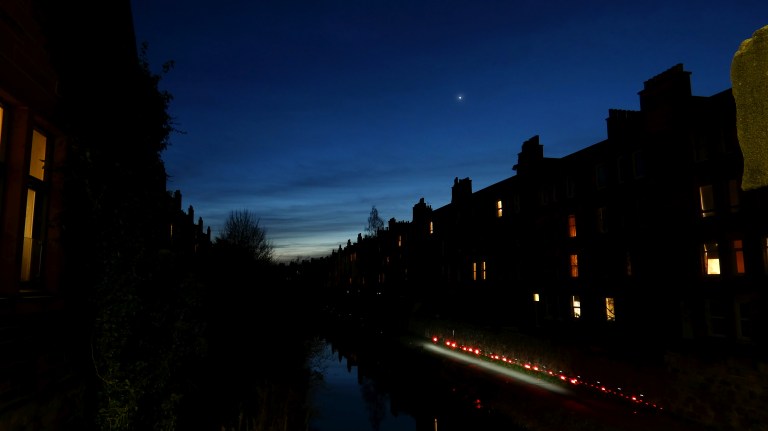 A twilight photograph taken along a narrow urban canal, with rows of silhouetted terraced buildings lining both sides and their lit windows glowing warm orange. Venus shines as a bright point of light in the deep blue evening sky above the center of the scene. A string of red pathway lights and a bright white light reflect off the still water of the canal below.