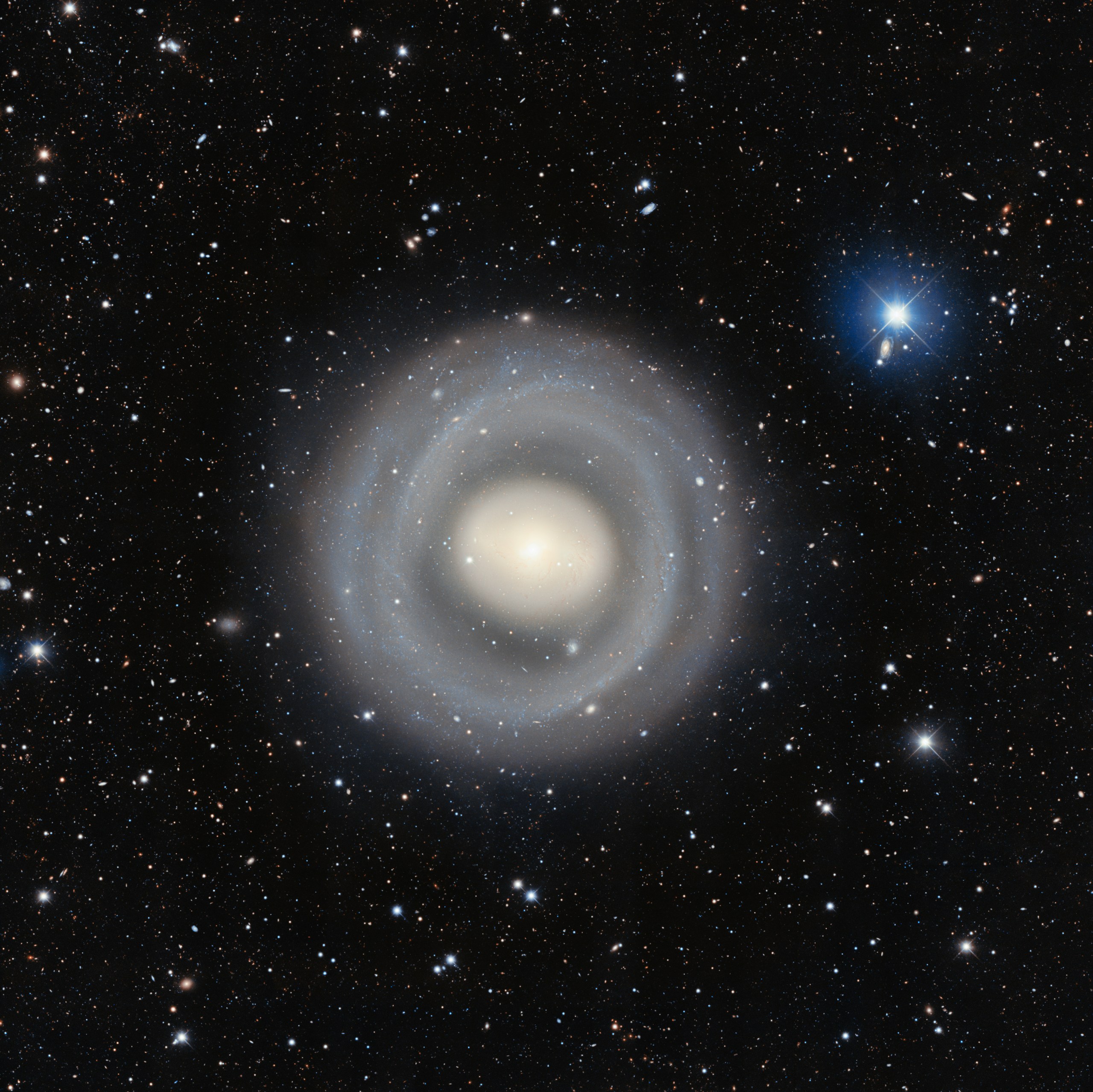 A telescopic photograph of a nearly face-on ringed spiral galaxy with a bright, glowing white-yellow core surrounded by multiple faint concentric blue-white spiral rings extending outward. The field is rich with distant background galaxies of various shapes and sizes scattered across the black sky, and a bright foreground star with prominent blue diffraction spikes is visible in the upper right.