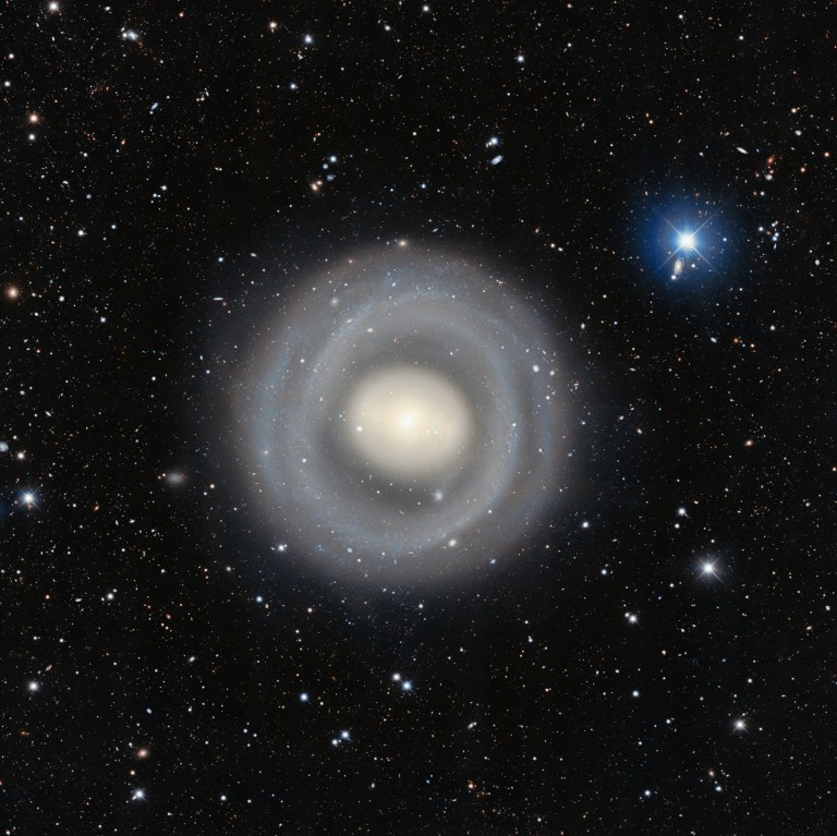 A telescopic photograph of a nearly face-on ringed spiral galaxy with a bright, glowing white-yellow core surrounded by multiple faint concentric blue-white spiral rings extending outward. The field is rich with distant background galaxies of various shapes and sizes scattered across the black sky, and a bright foreground star with prominent blue diffraction spikes is visible in the upper right.