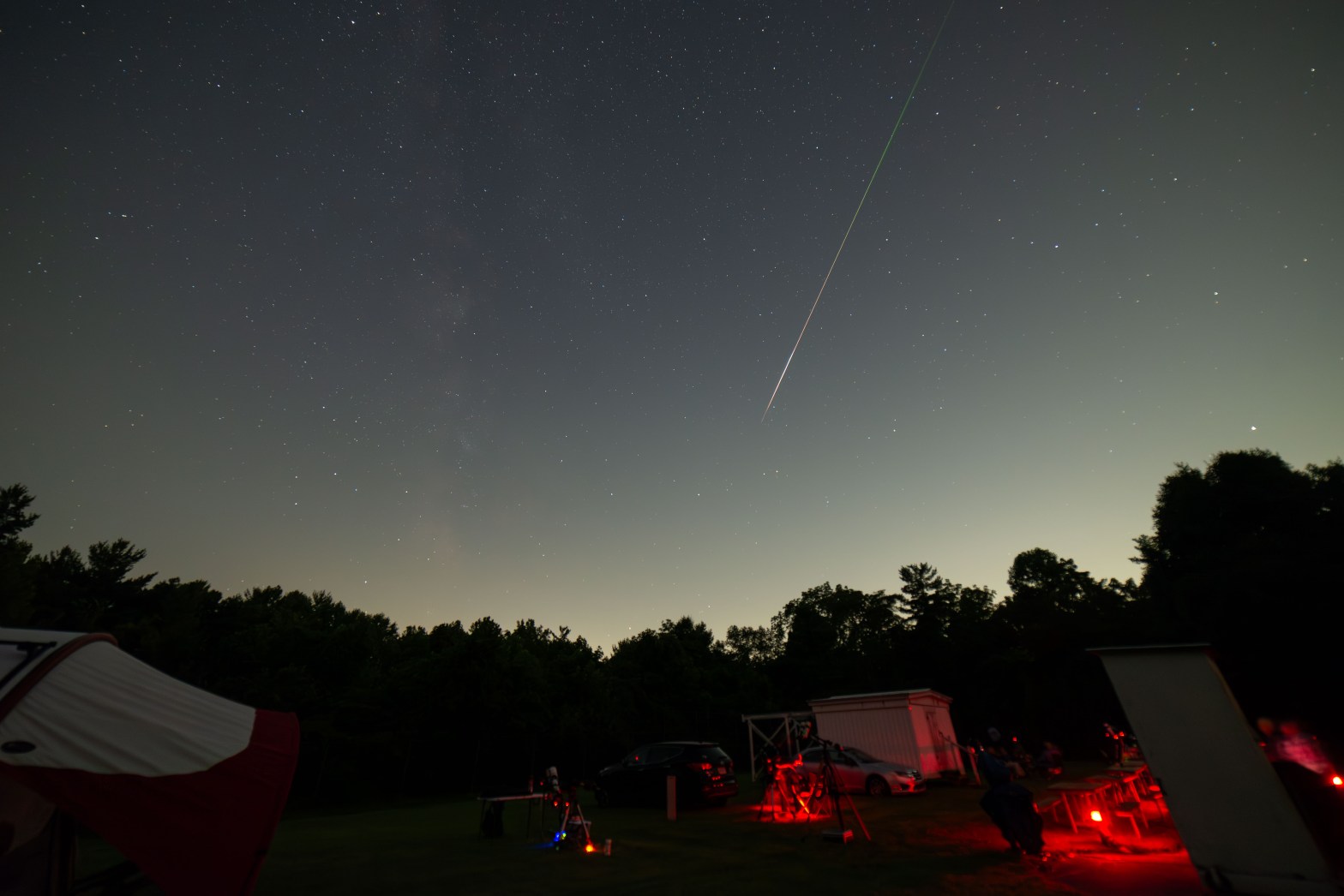 A wide-angle night sky photograph taken at a star party, showing a bright Perseid meteor streaking diagonally across the upper portion of the frame with a vivid green and white trail. The foreground shows an open field with several telescopes set up, observers lit by red lights to preserve night vision, cars, a small white building, and a treeline silhouetted against a faint twilight glow on the horizon. A red tent is partially visible in the lower left.