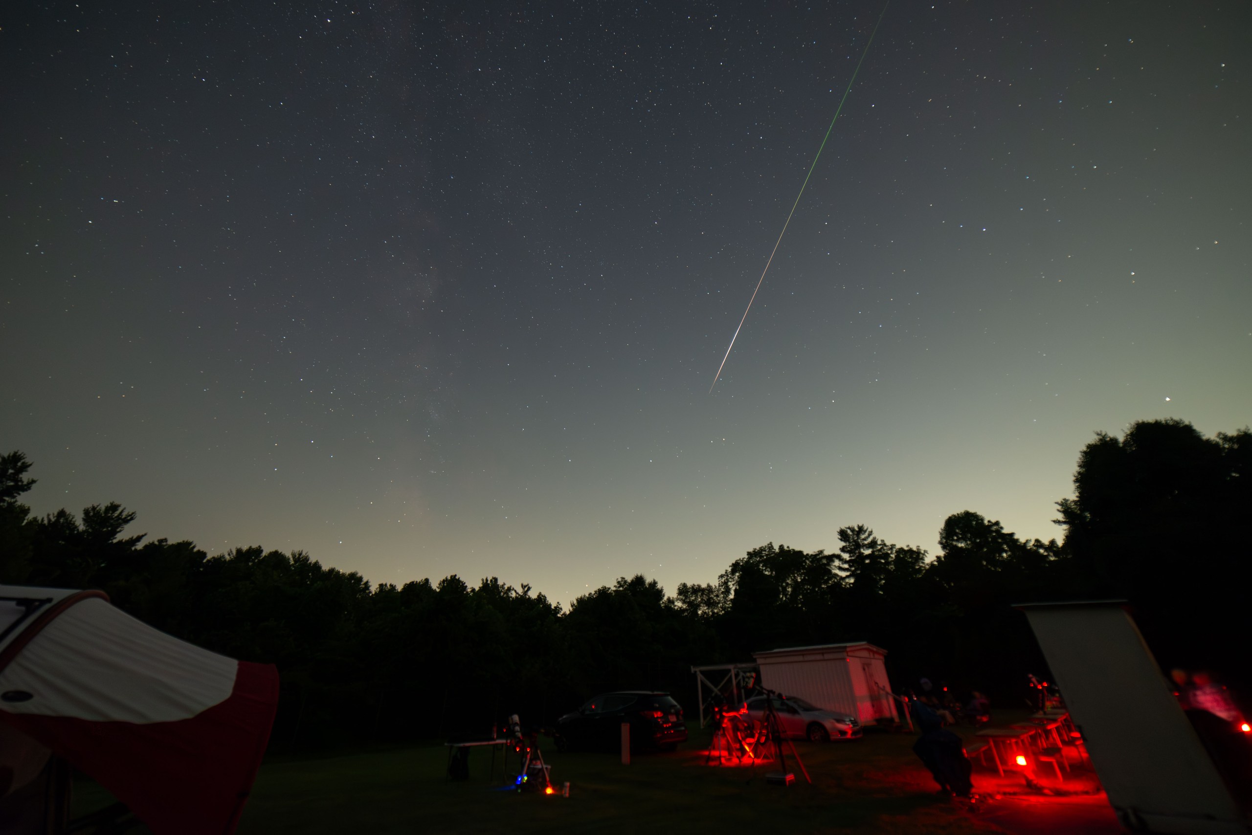 A wide-angle night sky photograph taken at a star party, showing a bright Perseid meteor streaking diagonally across the upper portion of the frame with a vivid green and white trail. The foreground shows an open field with several telescopes set up, observers lit by red lights to preserve night vision, cars, a small white building, and a treeline silhouetted against a faint twilight glow on the horizon. A red tent is partially visible in the lower left.