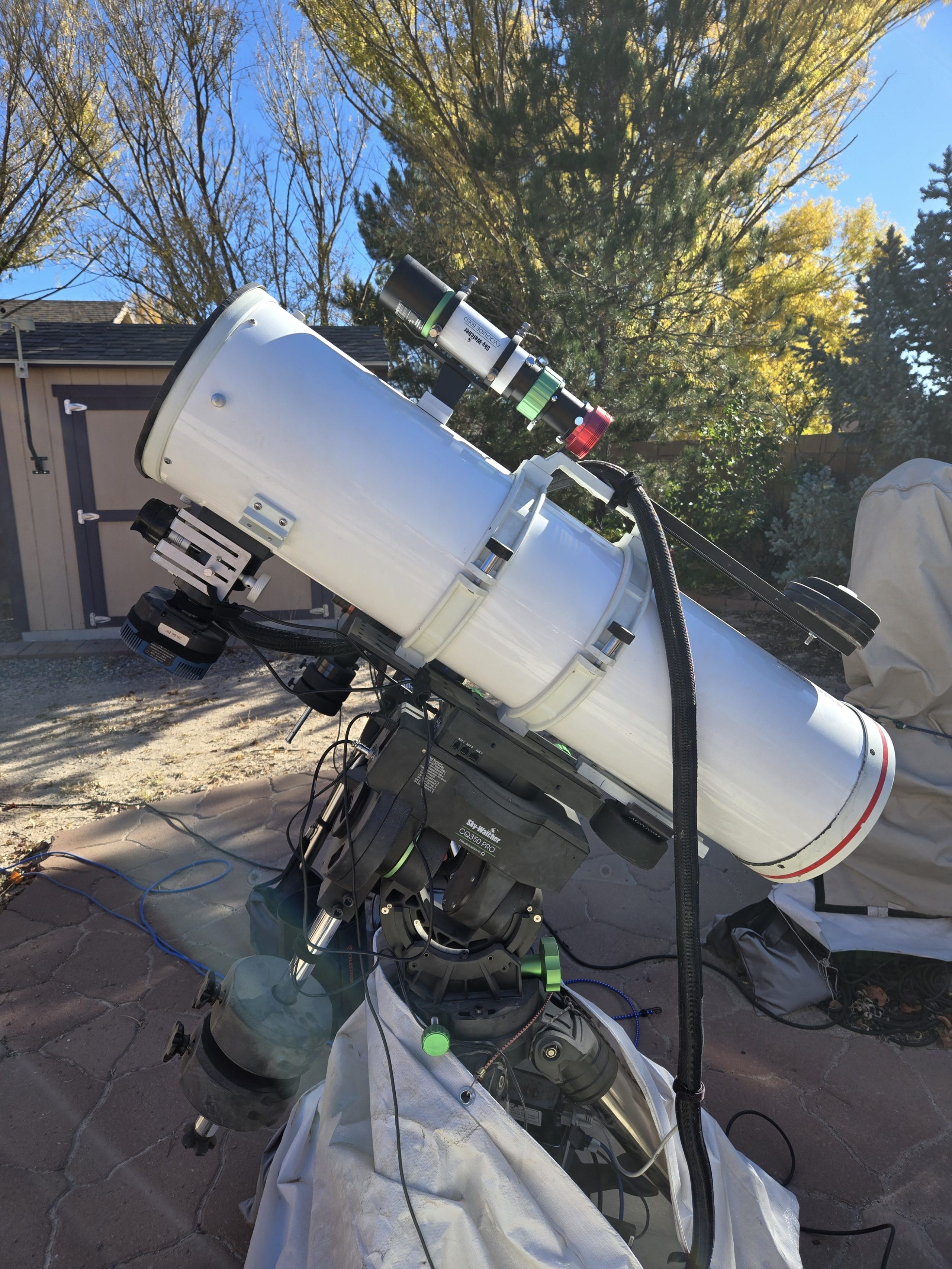 Photograph of a white Newtonian reflector telescope mounted on a black Sky-Watcher CQ350 Pro equatorial mount, sitting on a stone patio. A green-and-black finder scope is attached along the top of the main tube, with cables running from the mount, and the telescope is aimed upward toward the upper right. Bare and evergreen trees, a tan storage shed, and a blue sky fill the background, with a white cloth cover draped over equipment at lower right.
