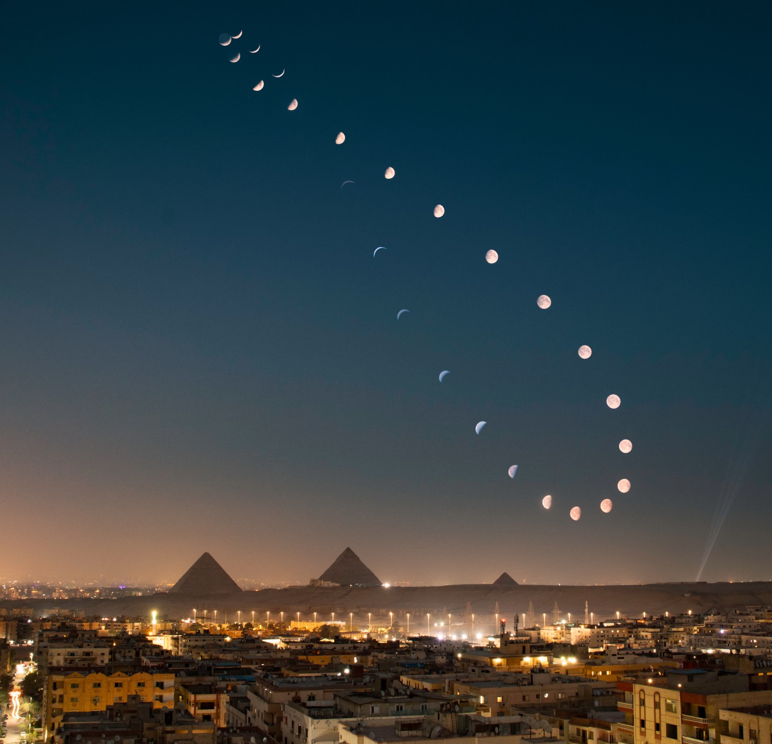 A composite photograph showing a lunar analemma — a sequence of Moon images captured at the same time over multiple nights — tracing a graceful figure-eight shaped arc across the night sky above the illuminated skyline of Giza, Egypt, with the three Great Pyramids silhouetted against the city's orange glow. The Moon's phases progress and change in size throughout the sequence, reflecting its varying distance and illumination over the course of the observation period.