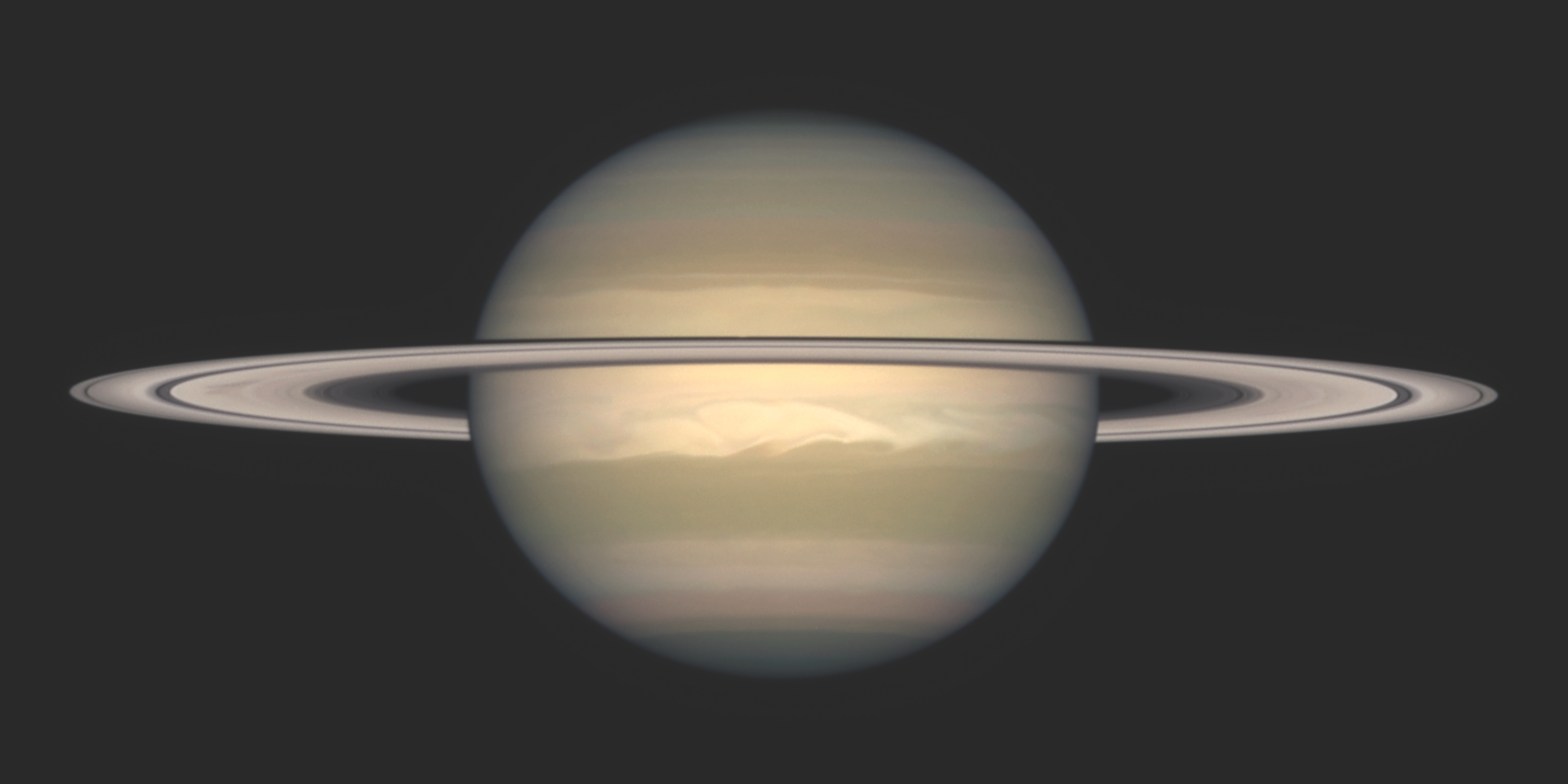 Photograph of Saturn against a dark background, seen nearly edge-on with its ring system stretching horizontally across the globe and casting a thin shadow band on the planet. The pale gold and tan cloud bands are visible, with a bright white storm feature swirling in the planet's southern hemisphere.