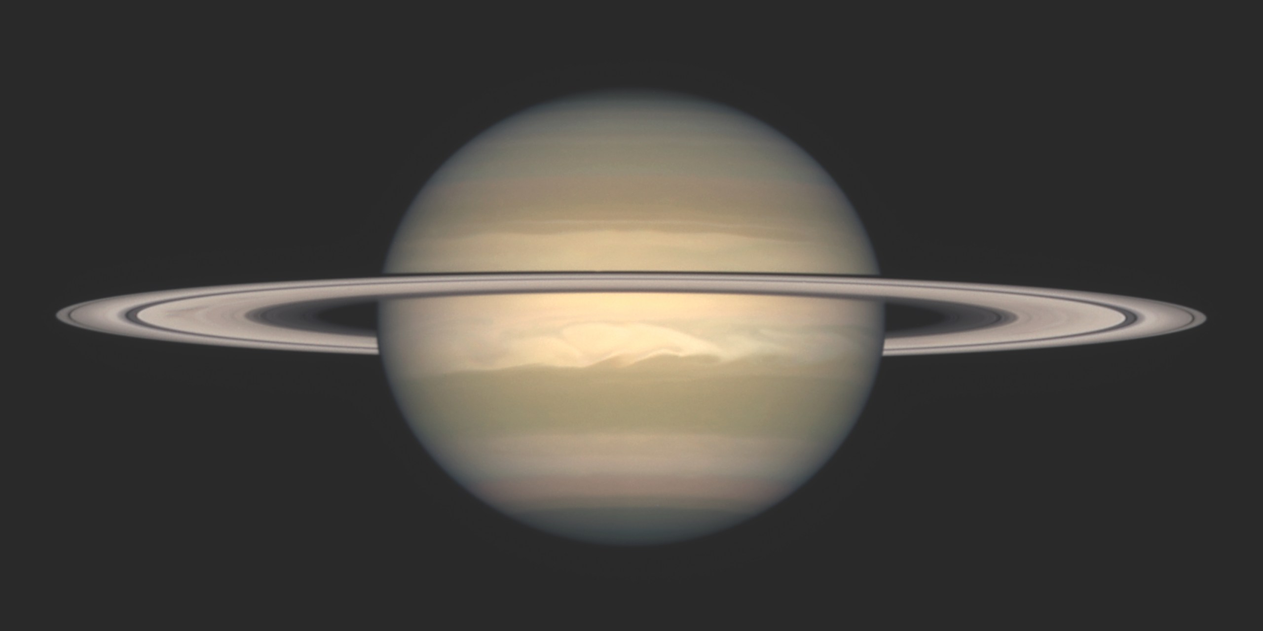 Photograph of Saturn against a dark background, seen nearly edge-on with its ring system stretching horizontally across the globe and casting a thin shadow band on the planet. The pale gold and tan cloud bands are visible, with a bright white storm feature swirling in the planet's southern hemisphere.