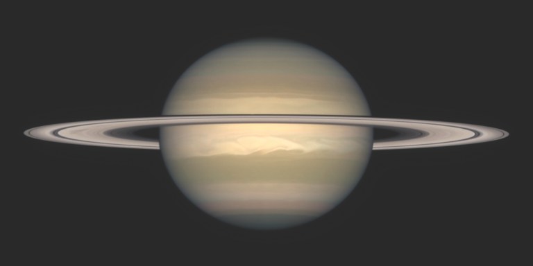Photograph of Saturn against a dark background, seen nearly edge-on with its ring system stretching horizontally across the globe and casting a thin shadow band on the planet. The pale gold and tan cloud bands are visible, with a bright white storm feature swirling in the planet's southern hemisphere.