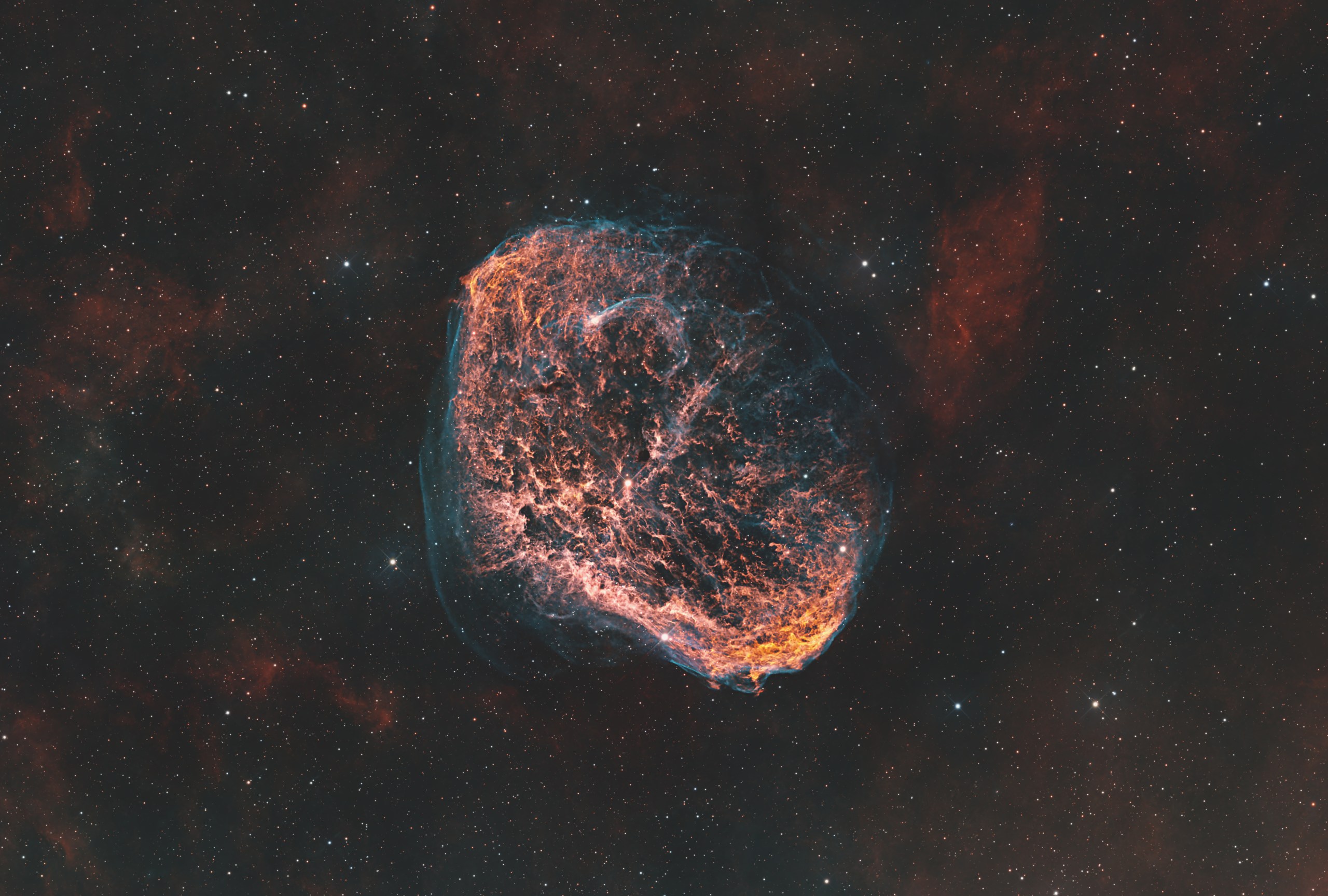A round, cloud-like shell fills the center of the frame, its interior a tangle of glowing orange and red wisps edged by a faint blue-green rim. The surrounding sky is dark and speckled with stars, with soft reddish haze drifting across the background.