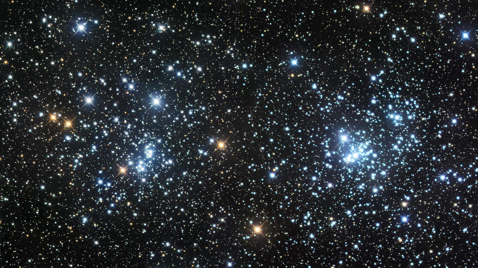 Photograph of the Double Cluster, showing two dense groupings of blue-white and orange stars side by side against a black sky thickly populated with fainter background stars, with the brightest stars in each cluster showing prominent diffraction spikes.