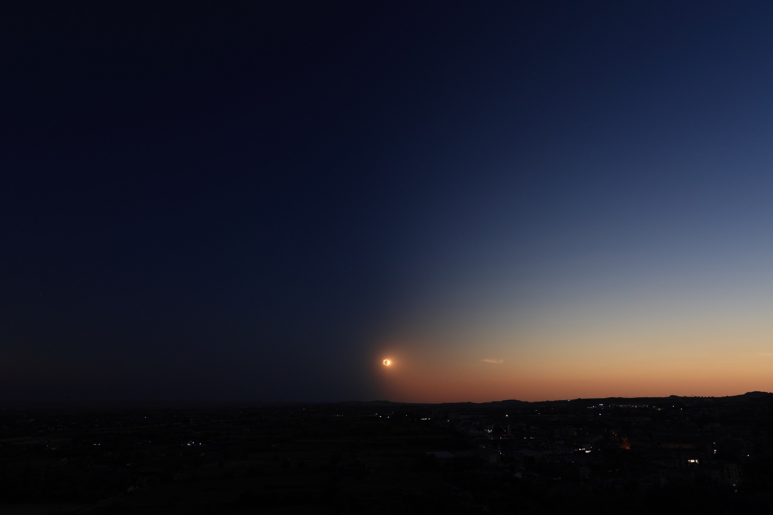 A vast sky fades from deep blue-black at the top to a warm orange glow near the horizon. Low in the sky, a small bright ring of light glows amber, like a thin crescent with a soft halo around it. Below, a dark, flat landscape stretches to the horizon, dotted with scattered pinpoints of town lights.