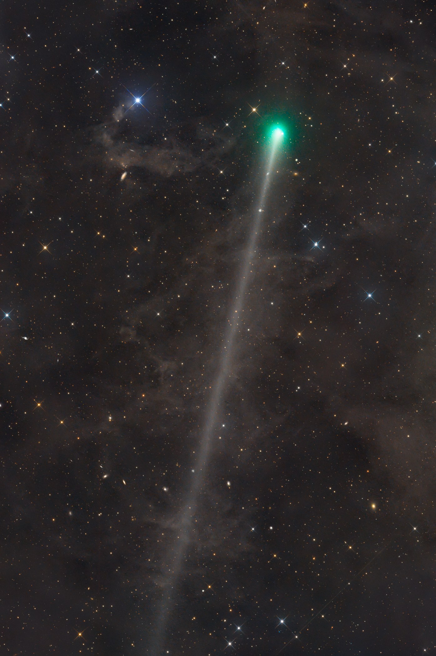 A bright green comet head glows near the top of a star-filled frame, streaming a long, faint white tail that stretches diagonally down almost the full height of the image. Wisps of dim brown dust clouds drift across the dark background, with a vivid blue-white star near the upper left.