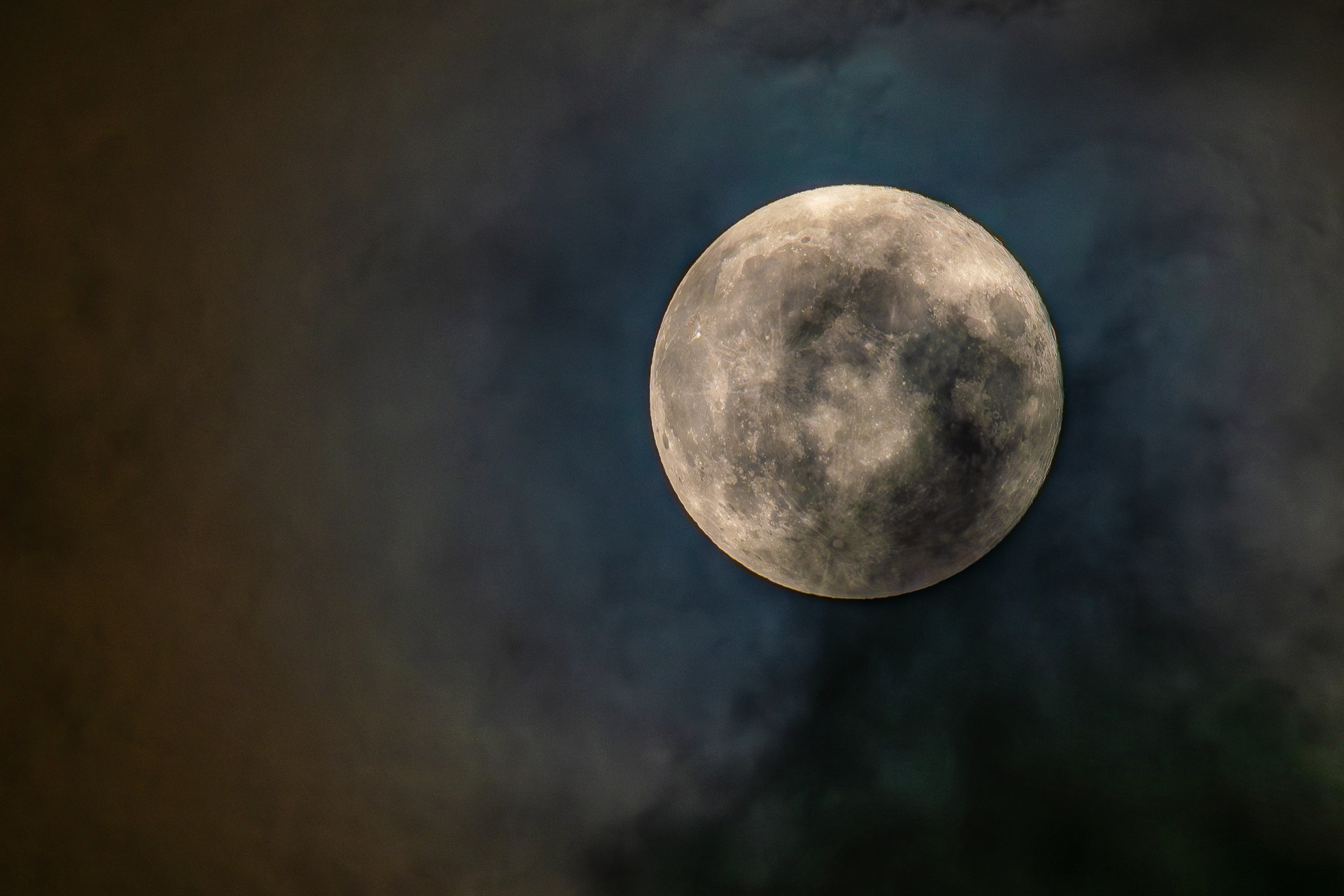 Photograph of a Full Moon showing detailed gray cratered terrain and dark mare regions, positioned right of center against a deep blue and amber sky streaked with soft clouds, with a darker cloud band trailing below the Moon.