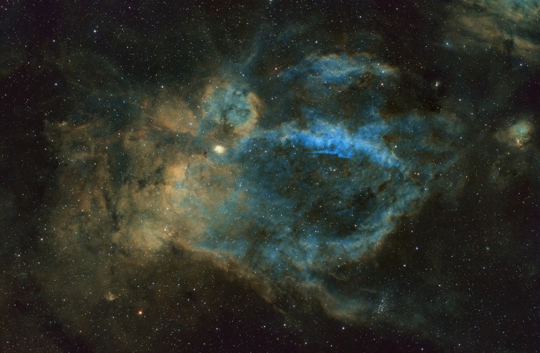 A cloud of green and gold gas fills most of the frame, with a bright blue oval structure stretching across the middle. Dark dust lanes thread through the brighter regions, and a small cluster of stars glows at the lower right.