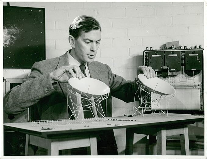 A black-and-white photograph shows a man in a suit jacket and tie leaning over a table, adjusting two small, white scale models of satellite dishes mounted on wire scaffolding. He looks down at the models with both hands resting lightly on the rims of the dishes. Behind him, a light-colored painted brick wall serves as the backdrop, flanked by a dark framed star map on the left and technical equipment with dials on the right.