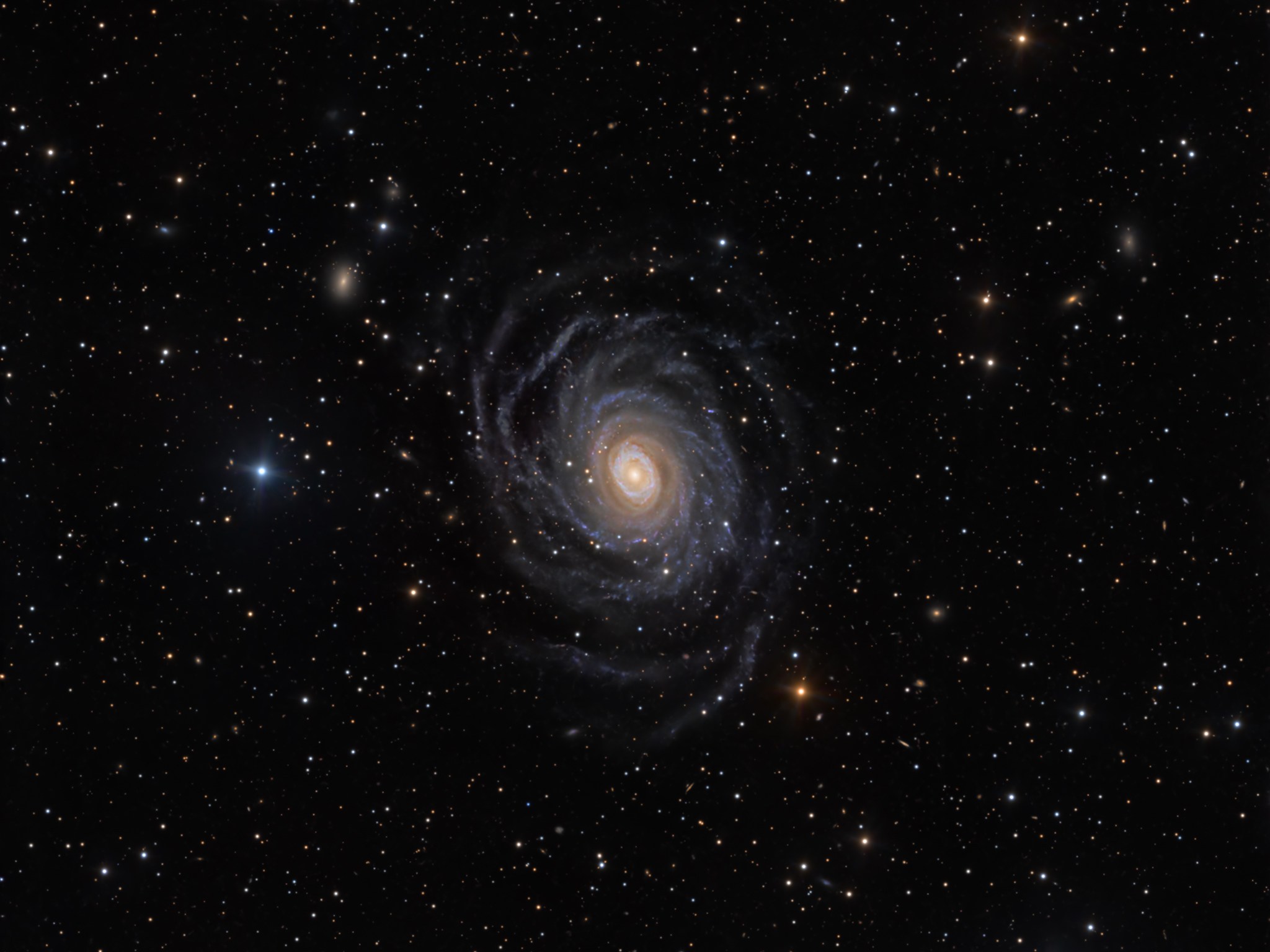 A galaxy with tightly wound, dusty blue spiral arms surrounds a glowing golden-yellow core, seen face-on against a dark, star-flecked background dotted with a few smaller, fainter galaxies. A bright blue-white star sits just to the left of the galaxy, and an orange star glows near the bottom edge of the frame.
