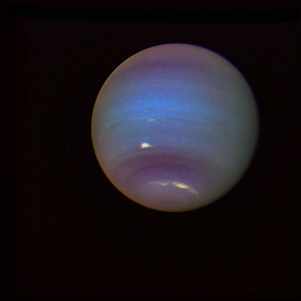 A Voyager 2 spacecraft image of Neptune showing the planet as a sphere with vivid blue and teal atmospheric bands, a large dark storm system visible in the southern hemisphere, and bright white clouds including a prominent high-altitude cloud streak near the center. The planet's limb glows with a subtle golden hue against the black background of space.