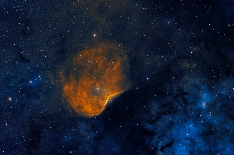A rounded, richly textured cloud of orange and gold gas fills the left side of the frame, its lower edge tapering to a bright point like a torch flame. Blue-tinted dust and haze spread across the right side, with faint dark filaments threading through a dense star field on a black background.