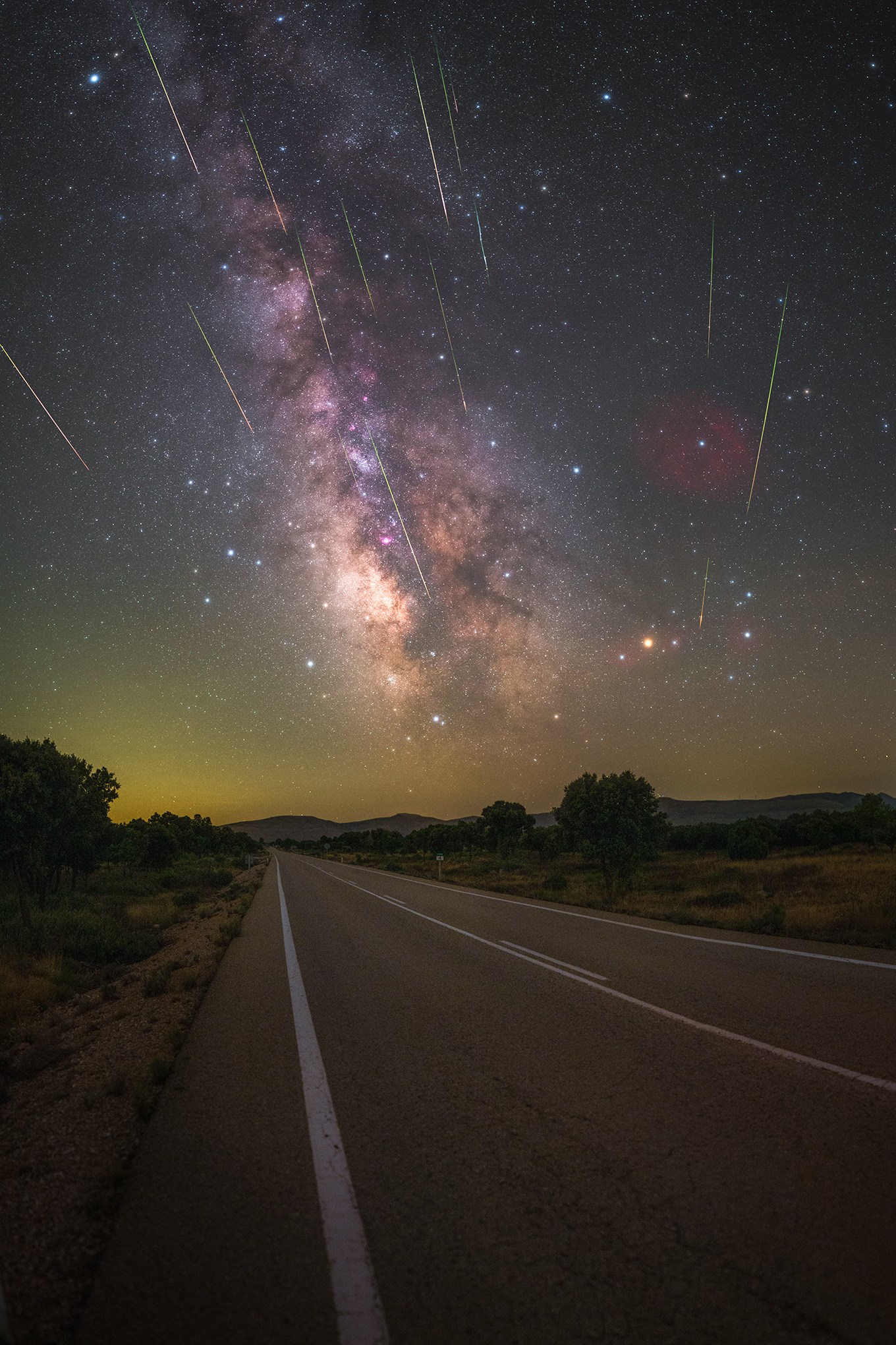 A straight two-lane road stretches toward a dark horizon, flanked by trees and grassy fields. Above it, the pale, textured band of the Milky Way arcs across the sky with a bright, pinkish glow at its core, while dozens of streaking meteors — white, green, and orange — shoot down through the stars in different directions. A faint yellowish glow lingers along the horizon.