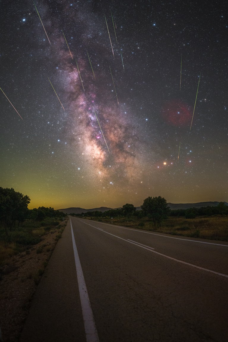 A straight two-lane road stretches toward a dark horizon, flanked by trees and grassy fields. Above it, the pale, textured band of the Milky Way arcs across the sky with a bright, pinkish glow at its core, while dozens of streaking meteors — white, green, and orange — shoot down through the stars in different directions. A faint yellowish glow lingers along the horizon.